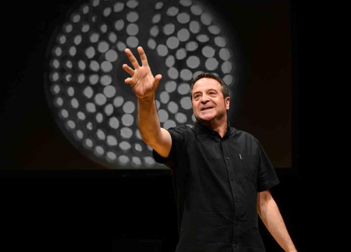 Man standing pointing forward with his right hand in front of a black screen with a white  circle design on it