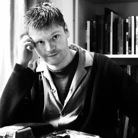 Black and white photograph of Rory Mullarkey looking to camera - at desk with bookshelves behind him