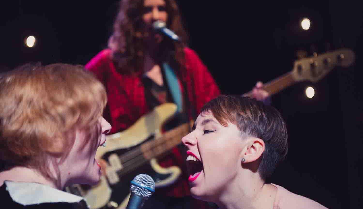 Photo of two women singing at each other with another woman playing the guitar in the background