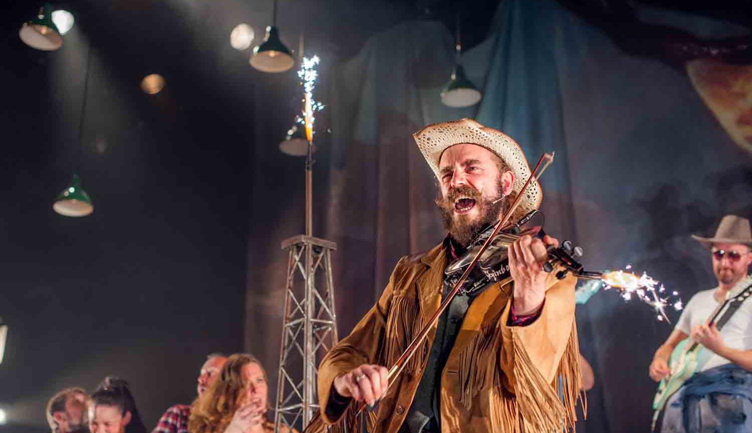 Photo of man with hat playing fiddle and singing on stage with other musicians in background