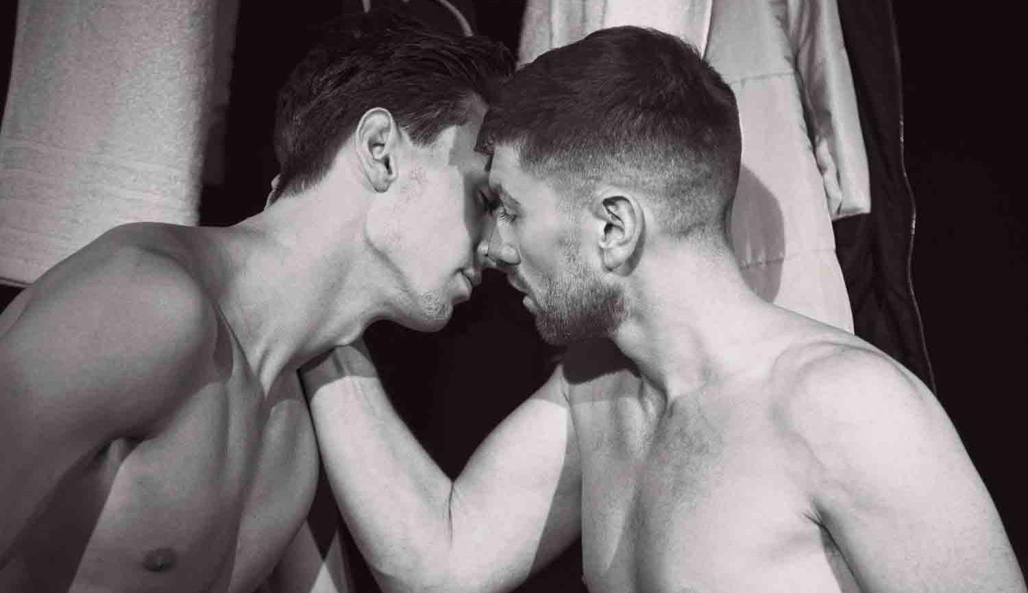 Black and white photo of two men with naked torsos kissing in a gym changing room