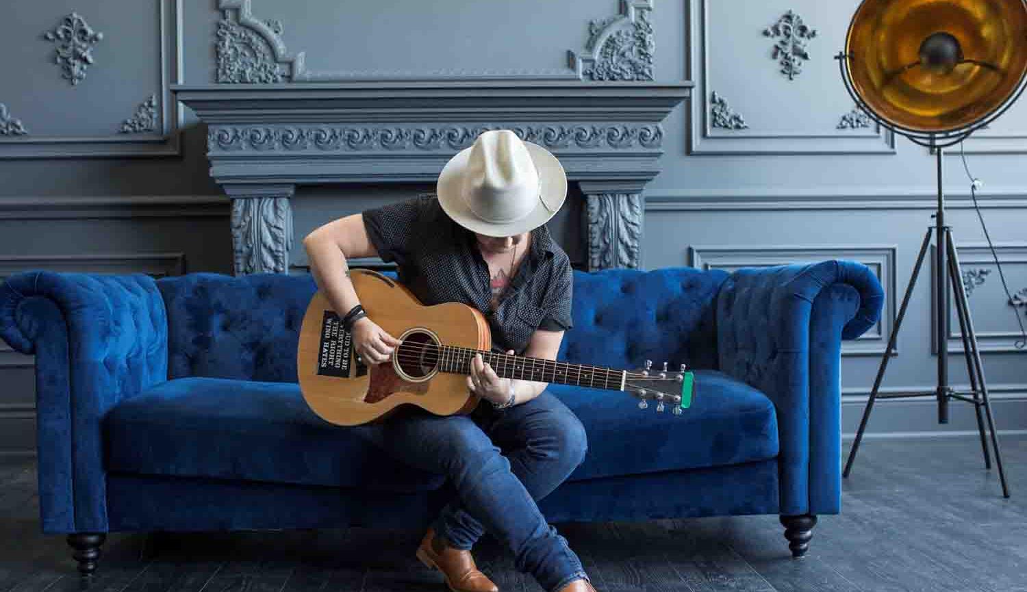 Photo of man sitting on sofa playing guitar wearing hat