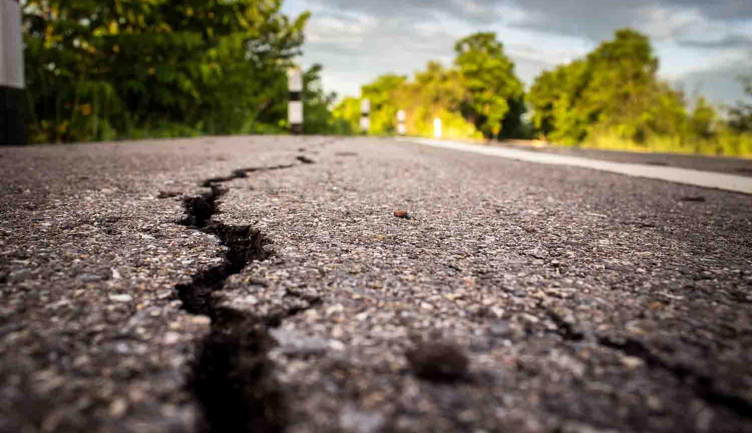 Photo at ground level of road with white line and crack in tarmac with trees in the distance