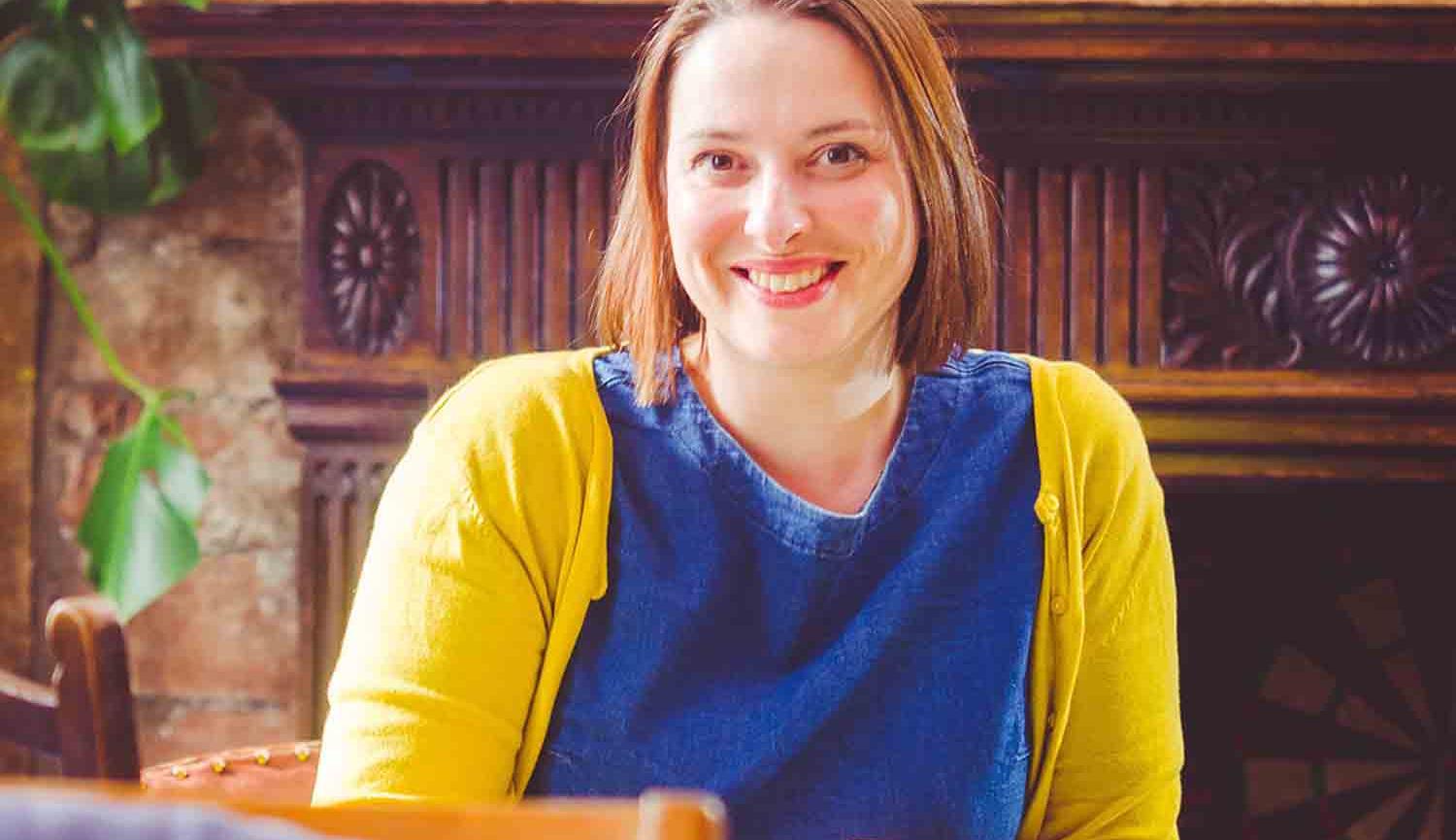 Photo of a woman sitting on a chair wearing a yellow cardigan and a blue top in front of a mantlepiece