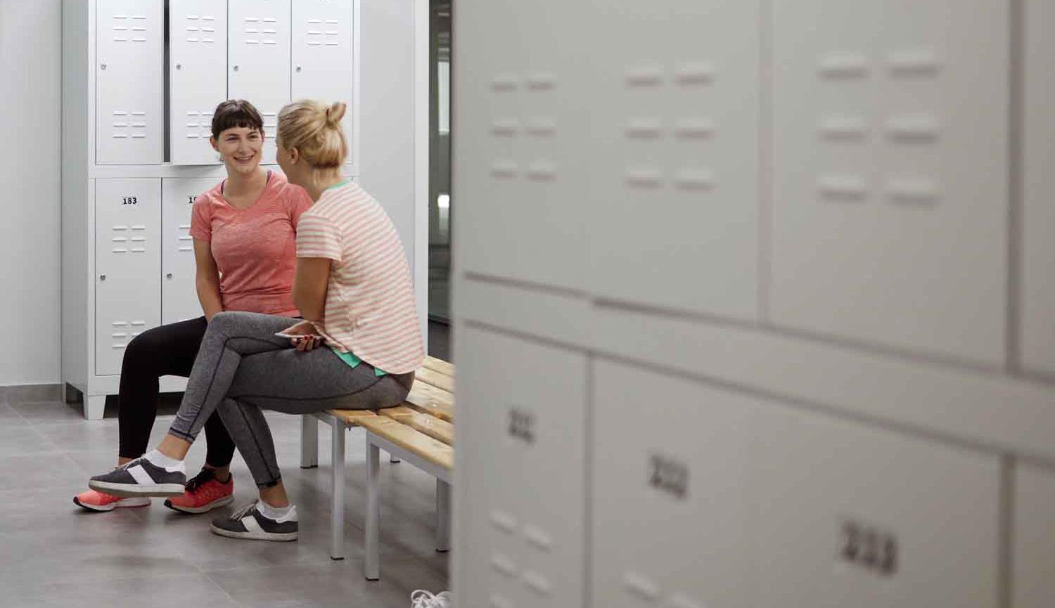 Photo of two women sitting  talking on bench in changing room with lockers around them