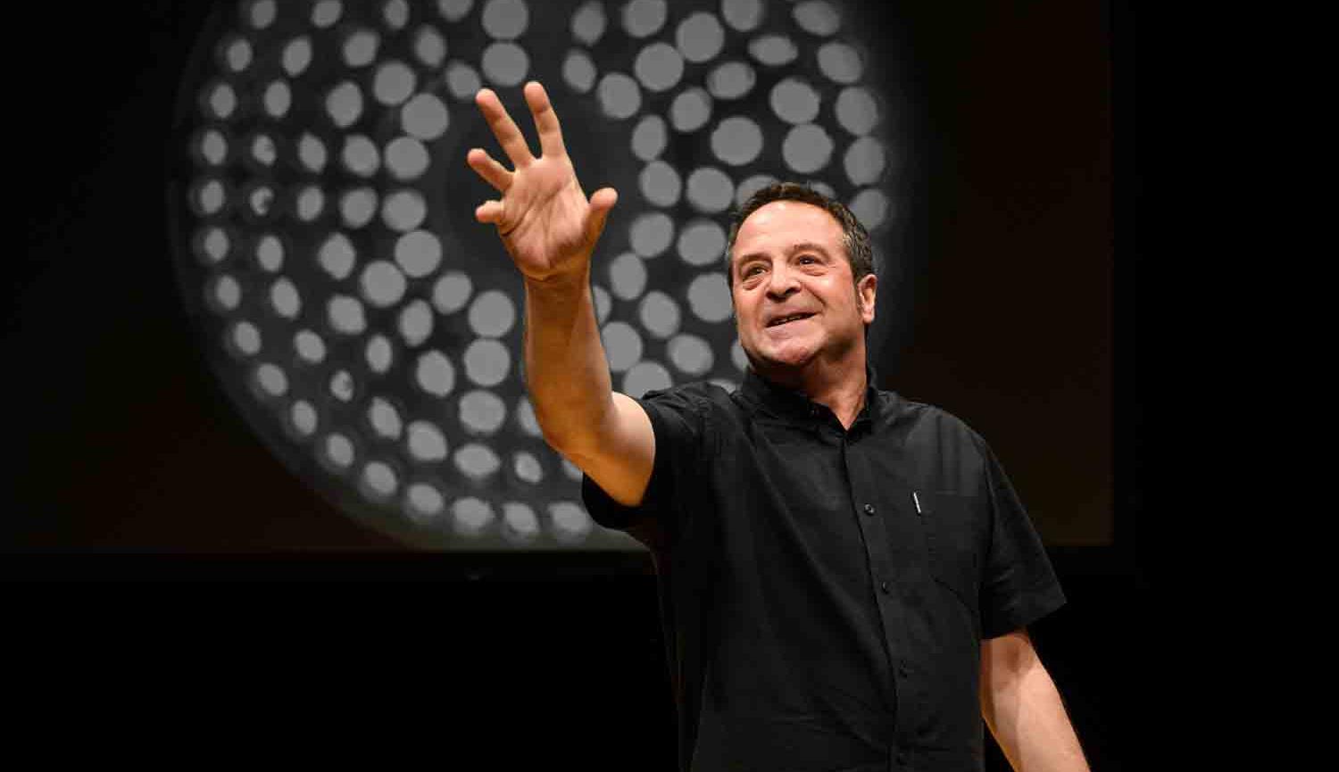 Photo of man pointing forwards with his right hand standing in front of black wall with white circle on it 