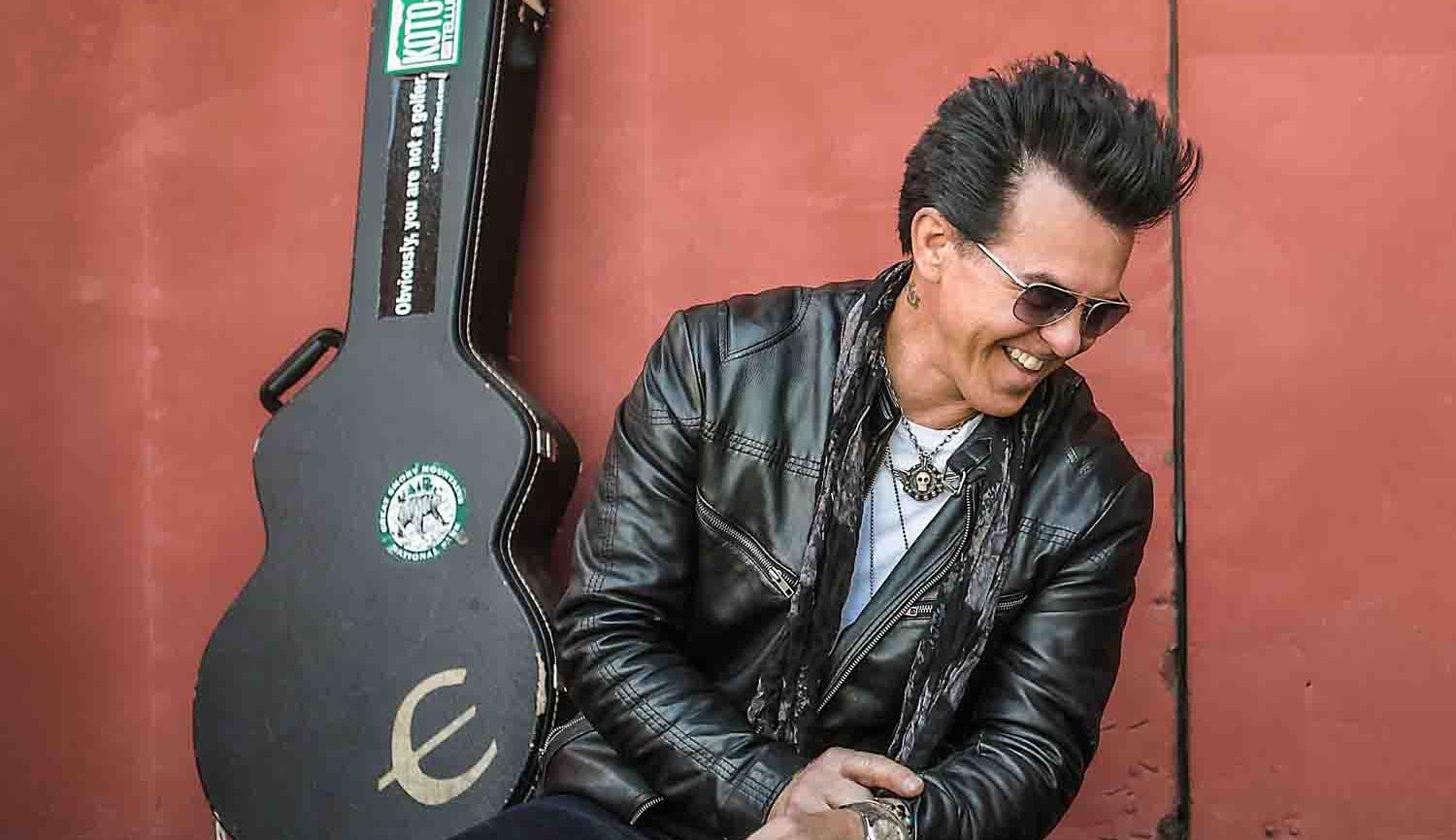 Photo of man sitting against a wall with a guitar case leaning against the wall behind him