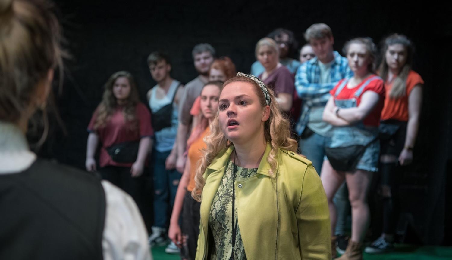 A young woman dressed in green speaks to someone else, their back is out of focus in the foreground