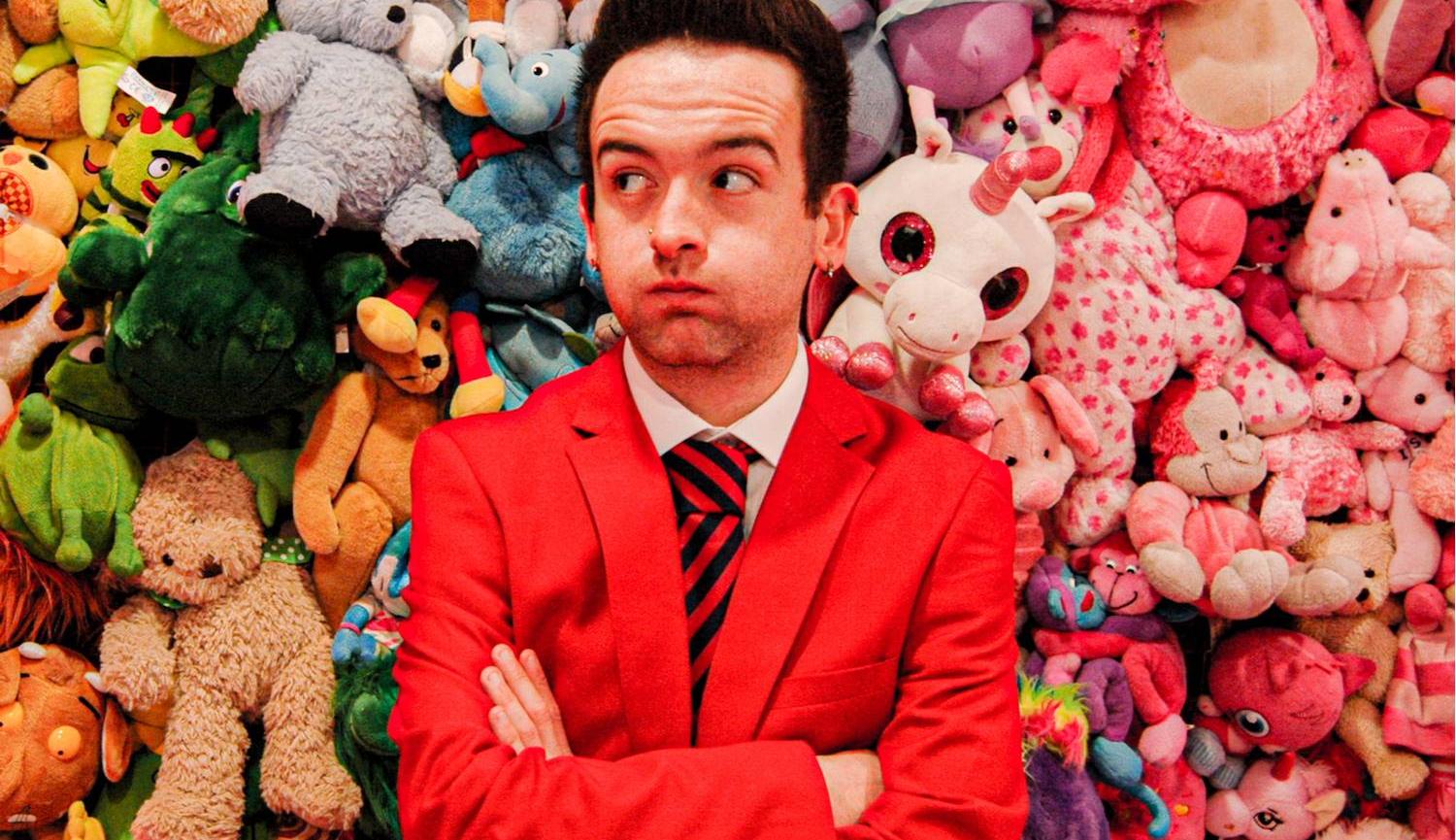 Lewis wearing his Redcoat jacket, standing with arms crossed, standing in front of a pile of soft toys