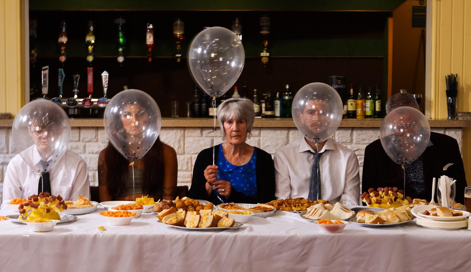 Five people sitting at a buffet table with balloons in front of their face