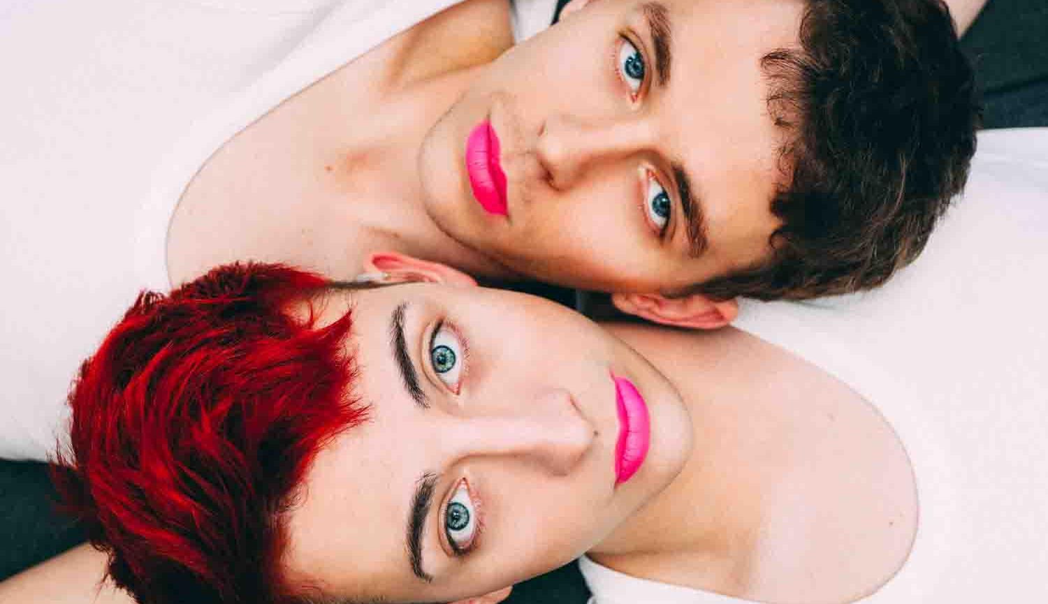 Head and shoulders photo of two men lying side by side wearing white vests and pink lipstick