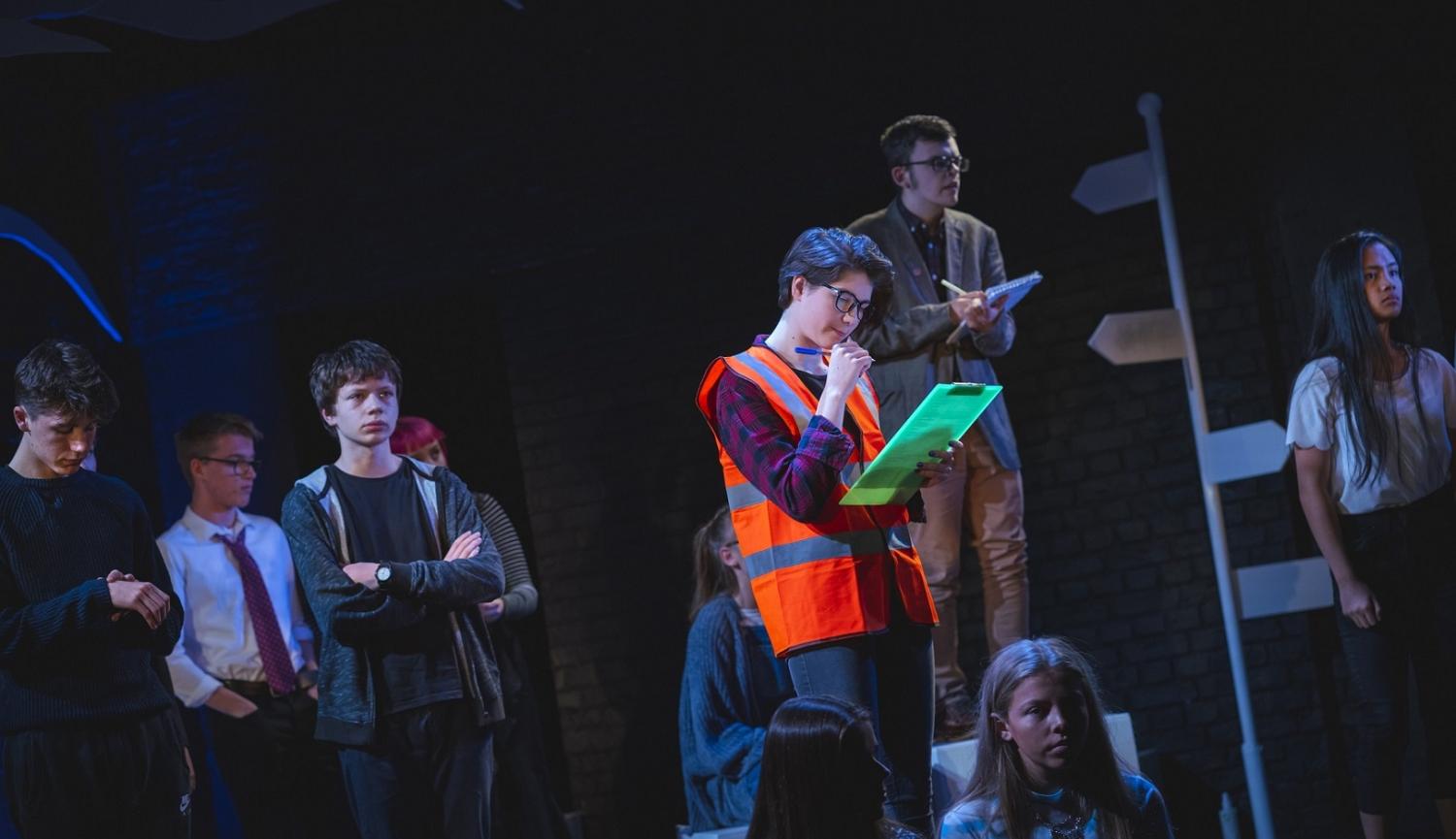 Photo of actors on stage with one wearing a high vis vest and holding a clipboard