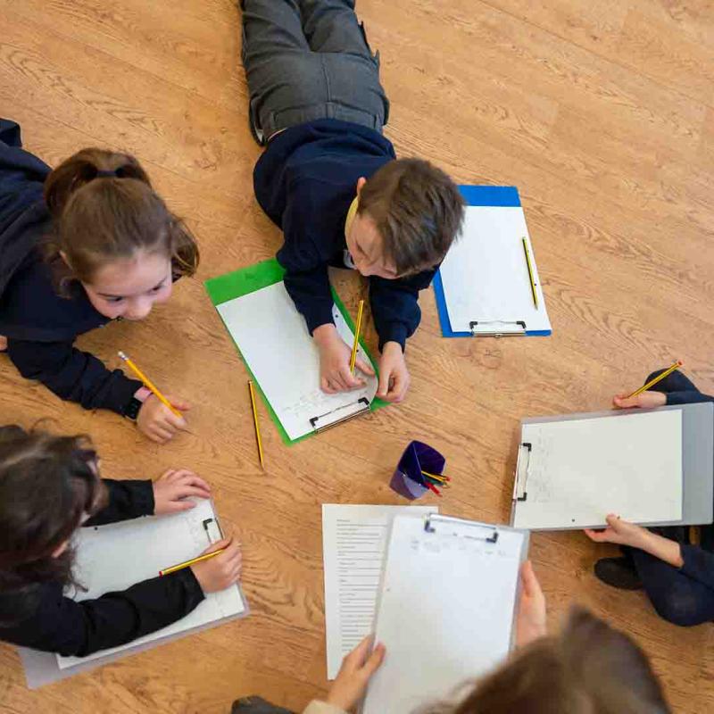 Group of school children lying on floor writing on paper on clipboards