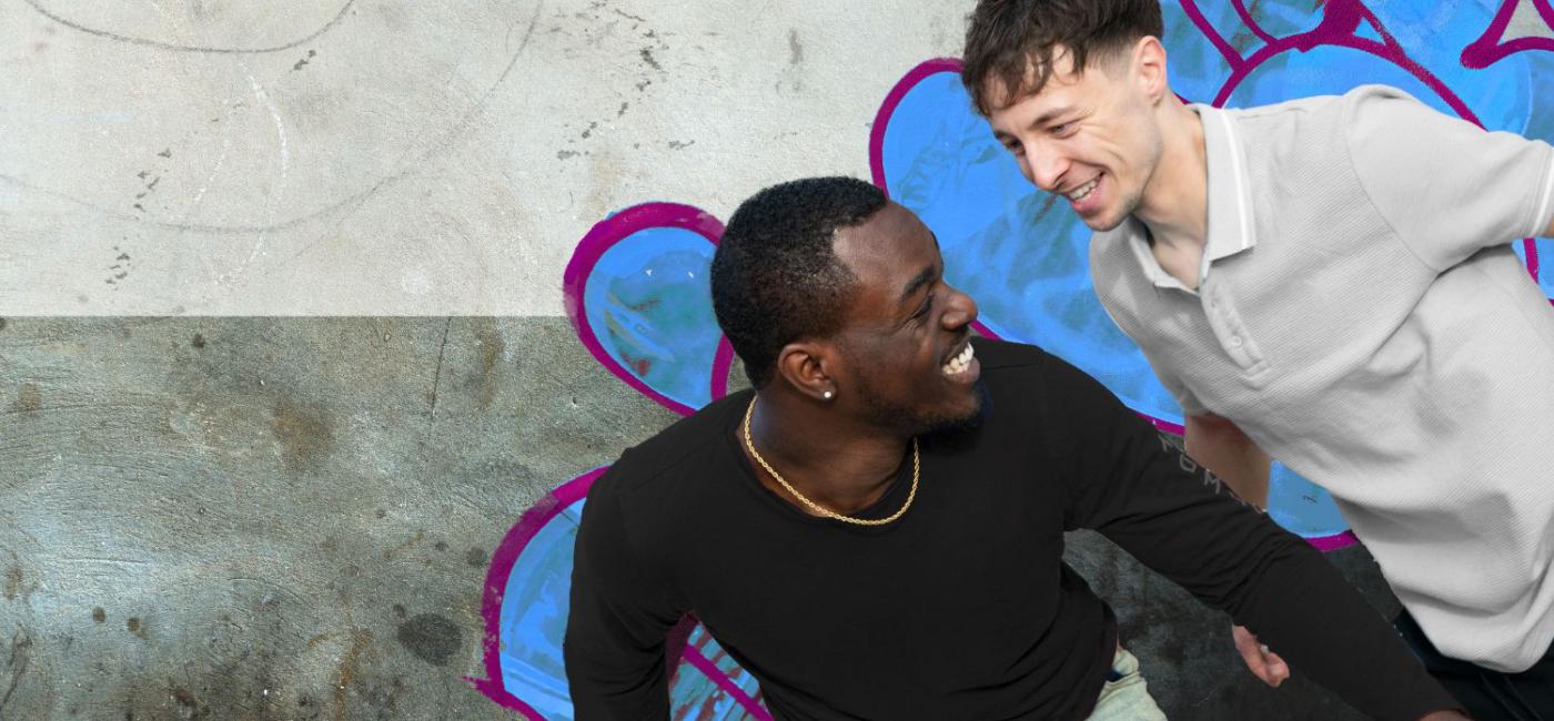 Two male friends stand in front of a graffiti covered wall, one black and one white