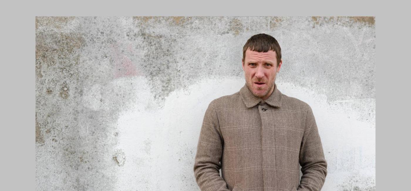 Jason Williamson standing in front of a grey wall with hands in his coat pockets