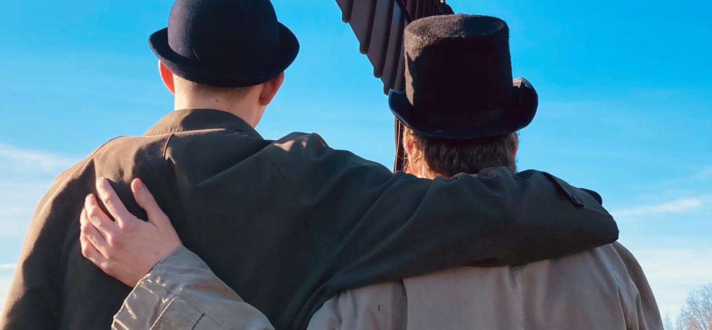 Two characters dressed in overcoats and hats stand in front of the Angel of the North