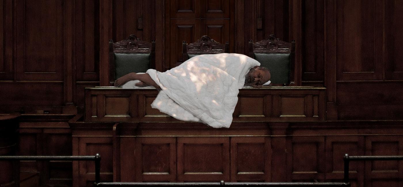 A person covered in a quilt lying on a wooden bench in a courtroom
