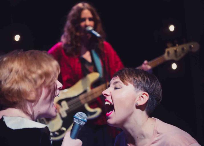 Photo of two women singing at each other with another woman playing the guitar in the background