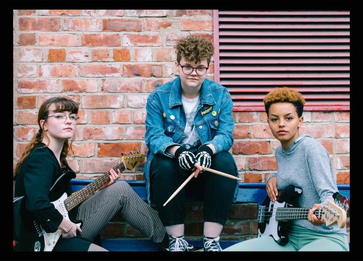 Cast members Elena, Bethany and Bridget sitting on a bench with guitars and drumsticks