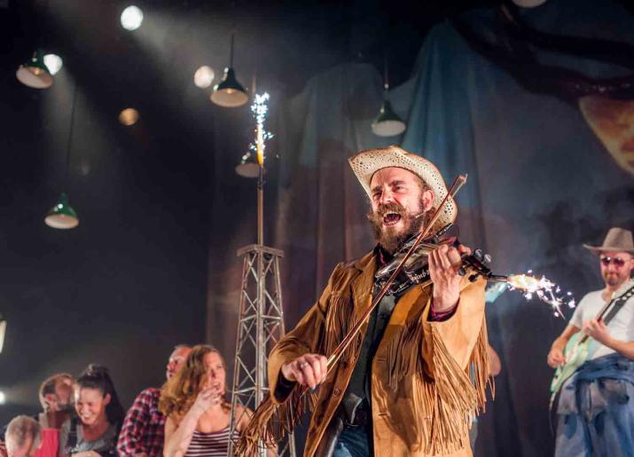 Photo of man with hat playing fiddle and singing on stage with other musicians in background