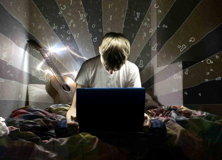 Photo of boy sitting on bed looking at laptop with guitar behind him