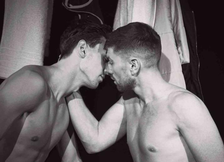Black and white photo of two men with naked torsos kissing in a gym changing room