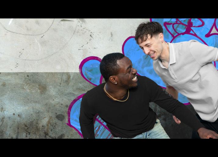 Two male friends stand in front of a graffiti covered wall, one black and one white