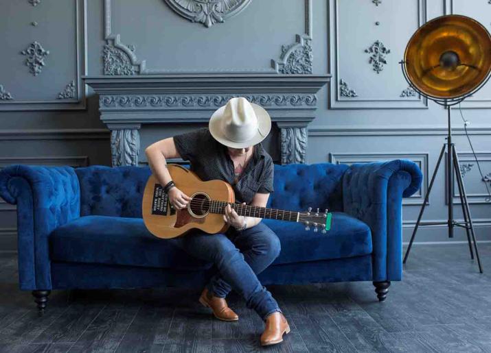 Photo of man sitting on sofa playing guitar wearing hat