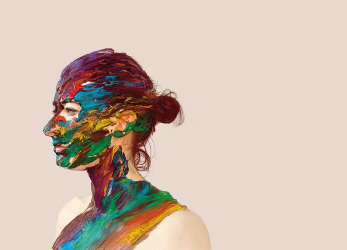 Photo of head and shoulders of woman standing side on to camera with lots of different colours of paint on her skin and hair