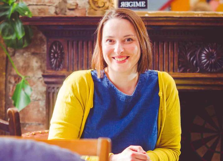 Photo of a woman sitting on a chair wearing a yellow cardigan and a blue top in front of a mantlepiece