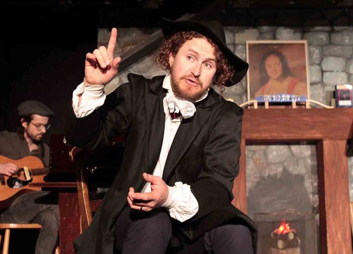 Photo of man in black hat, black jacket and white shirt sitting down, pointing one finger on one hand upwards