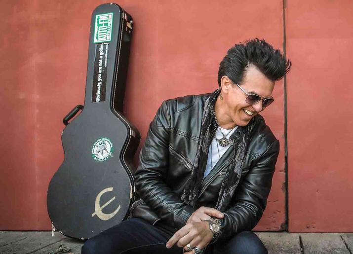 Photo of man sitting against a wall with a guitar case leaning against the wall behind him