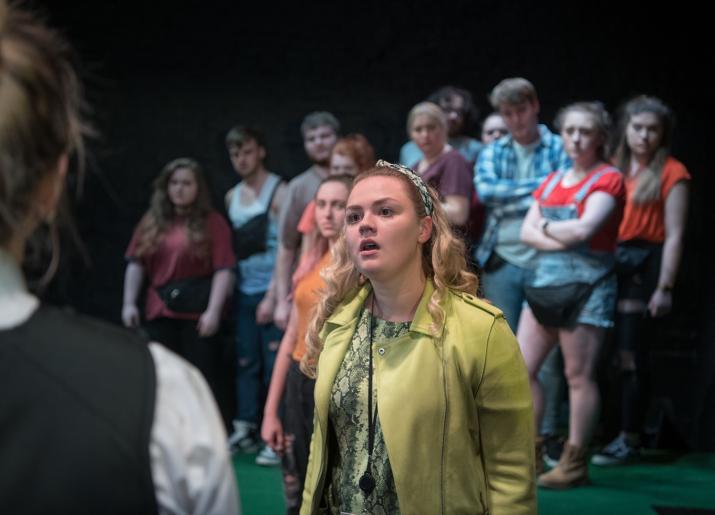 A young woman dressed in green speaks to someone else, their back is out of focus in the foreground