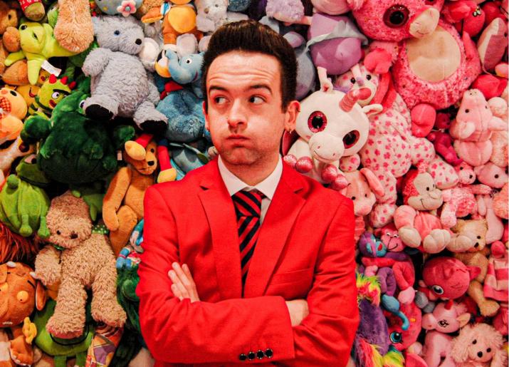 Lewis wearing his Redcoat jacket, standing with arms crossed, standing in front of a pile of soft toys