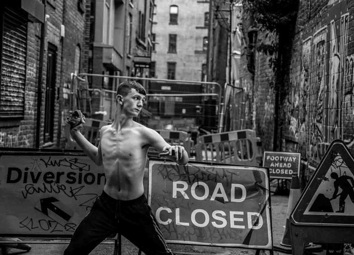 Black and white photo of young man about to throw a brick, with a naked torso, wearing trousers, standing in front of a derelict alley with road closed signs behind him