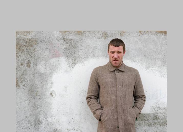 Jason Williamson standing in front of a grey wall with hands in his coat pockets