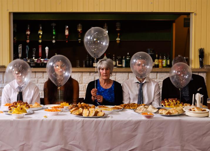 Five people sitting at a buffet table with balloons in front of their face