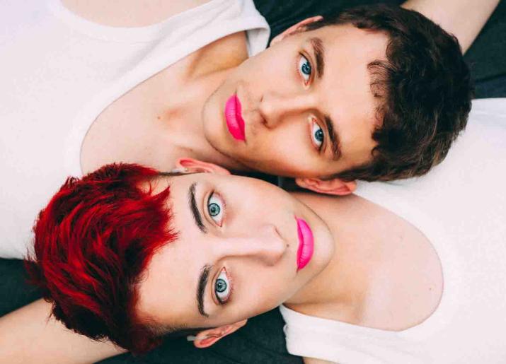 Head and shoulders photo of two men lying side by side wearing white vests and pink lipstick