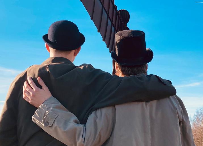 Two characters dressed in overcoats and hats stand in front of the Angel of the North