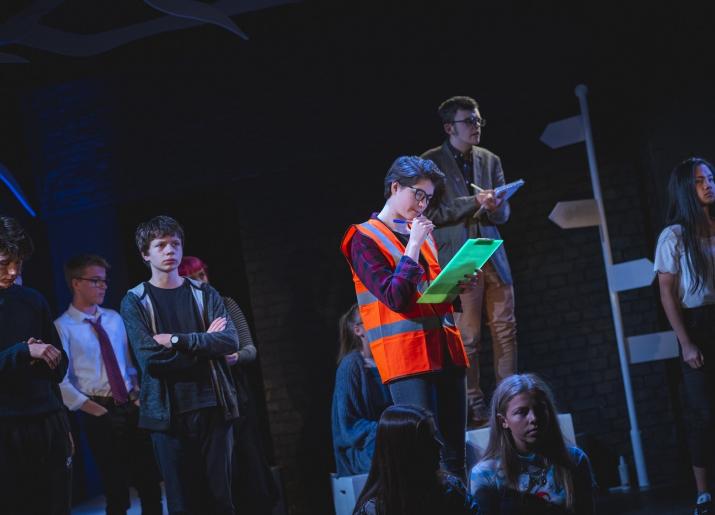 Photo of actors on stage with one wearing a high vis vest and holding a clipboard