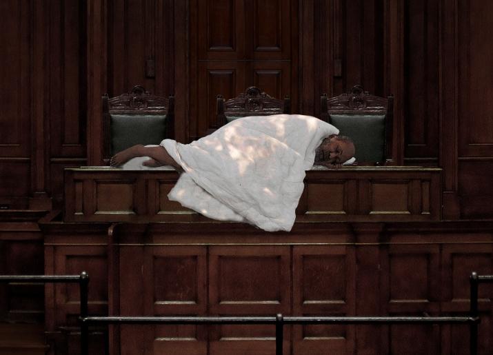 A person covered in a quilt lying on a wooden bench in a courtroom