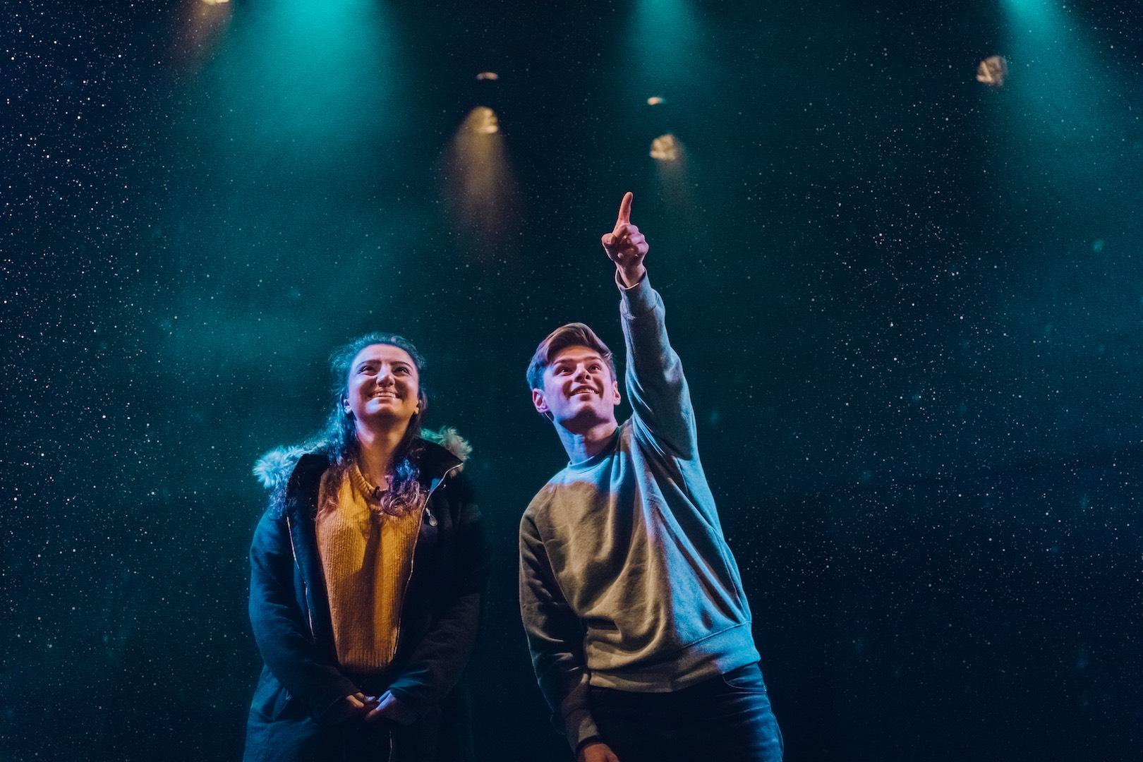 A young lady and young man on stage - the man is pointing to something high up in the distance