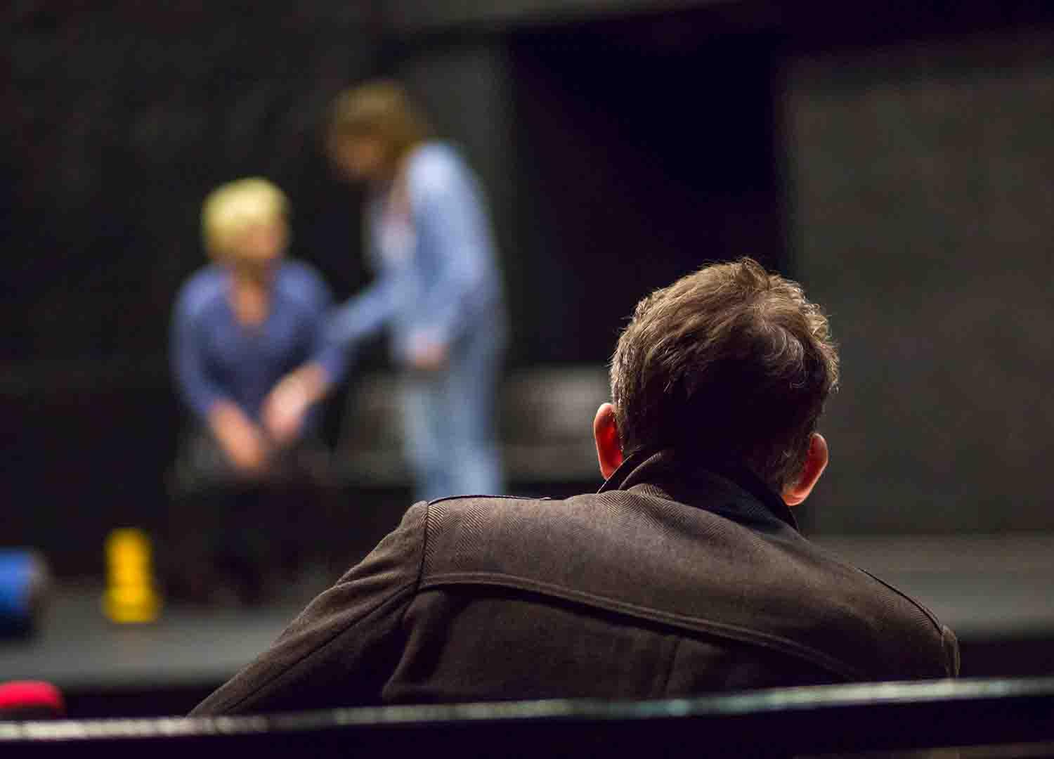 Photo of back of mans head and shoulders sitting watching two people on stage